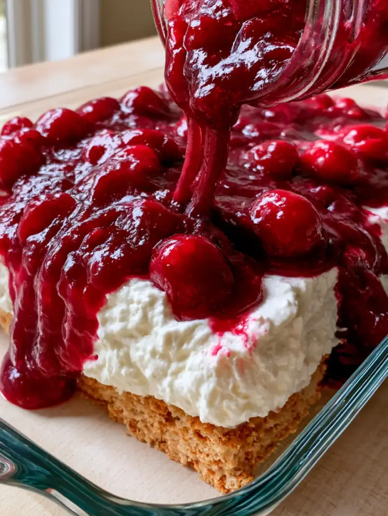 Wide shot of the cherry cream cheese dessert in a glass dish on a light wood countertop with natural sunlight highlights.