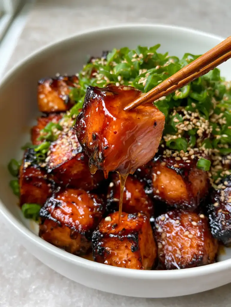 A wooden chopstick lifting a single dripping, glistening honey garlic salmon bite showing the juicy pink interior.